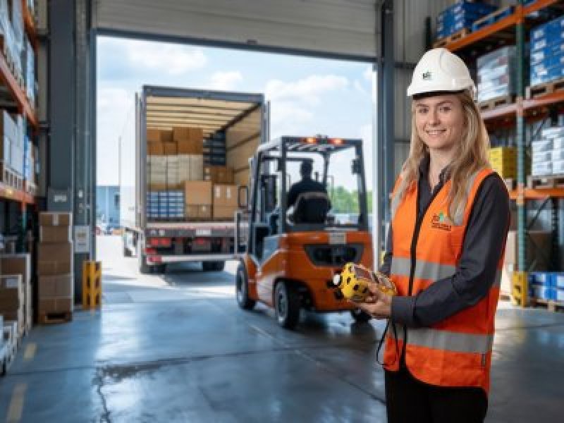 JTA consultant conducting on-site workplace safety inspection in a warehouse environment. JTA field consultant wearing a white hard hat and orange high-visibility vest stands inside a warehouse, holding a gas detection device while a forklift loads a truck in the background.