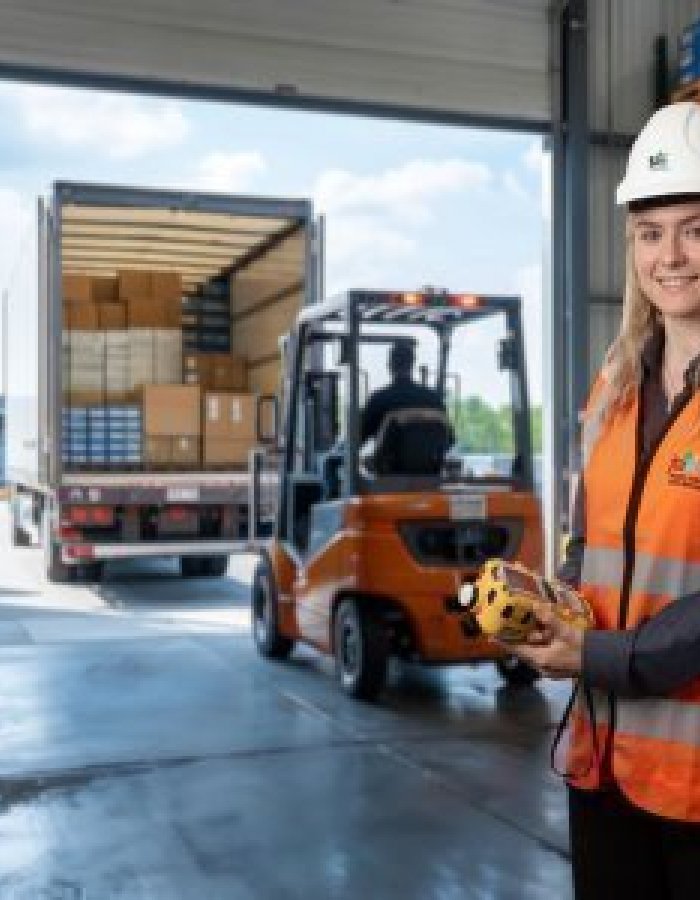 JTA field consultant wearing a white hard hat and orange high-visibility vest stands inside a warehouse, holding a gas detection device while a forklift loads a truck in the background.
